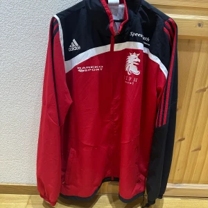 Röd och svart träningsjacka från Adidas - Säljer en sportig röd och svart träningsjacka från Adidas med vita detaljer och klubbmärke på bröstet. Jackan har dragkedja, lång ärm och två fickor framtill. Perfekt för fotbollsträning eller som snygg streetwear. Helt oanvänd. 