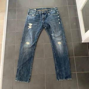 Blå slitna jeans från Altatensione - Säljer ett par blå jeans från Altatensione med slitna detaljer och raka ben. Jeansen har klassisk femficksdesign, gylf med dragkedja och en liten metallogo på bakfickan. Perfekta för en avslappnad stil. Det finns ingen lapp med storleken på, men enligt mig sitter de som storlek 32/32. Jag kan skicka mått om det skulle vara aktuellt.