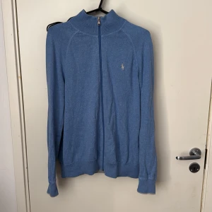 Blå stickad kofta från Ralph Lauren - Säljer en blå stickad kofta från Ralph Lauren med dragkedja framtill och broderad logga på bröstet. Tröjan har lång ärm och hög krage, perfekt för lager-på-lager. Klassisk och stilren modell.