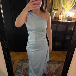 Ljusblå långklänning med oneshoulder - Säljer en elegant ljusblå långklänning med oneshoulder och draperad detalj i midjan samt en slits. Klänningen har en smickrande passform och är tillverkad i ett glansigt material som ger en lyxig känsla. Perfekt för speciella tillfällen. Säljer för att jag beställde två i samma färg och valde den andra. Kom privat för fler bilder💕