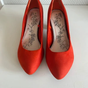 Röd/ orangea pumps med klack - Snygga röda/ orangea pumps från Vox med spetsig tå och klack. Skorna är i mocka och har en stilren design med blommigt mönster på innersulan. Perfekta för dig som vill sticka ut med färg.