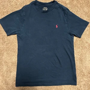 Svart t-shirt från Polo Ralph Lauren - Säljer en klassisk mörkblå t-shirt från Polo Ralph Lauren med röd broderad logga på bröstet. T-shirten har rund halsringning och korta ärmar. Perfekt för en stilren och avslappnad look.
