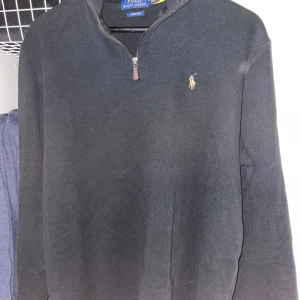 Svart halvzip tröja från Polo Ralph Lauren - Klassisk svart långärmad half zip från Ralph Lauren med logga på bröstet. Tröjan har ribbade muddar och är perfekt för en stilren och avslappnad look.