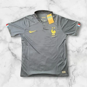 Frankrike fotbollströja Nike grå - Säljer en grå Frankrike fotbollströja från Nike med gula detaljer och emblem på bröstet. Tröjan har korta ärmar och en klassisk krage med knapp. Franska flaggans färger syns på ärmsluten. Perfekt för fans av landslaget!