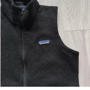 Svart fleeceväst från Patagonia - Svart fleeceväst från Patagonia med dragkedja framtill och ståkrage. Västen har två fickor med dragkedja och en liten logotyp på bröstet. Perfekt för lager-på-lager och enkel att matcha.