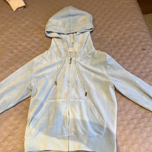 Ljusblå velour hoodie från Juicy Couture - Säljer en ljusblå hoodie i mjuk velour från Juicy Couture. Tröjan har dragkedja, huva med snören och fickor framtill. Broderad JC-logga på bröstet.