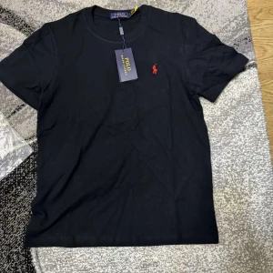 Svart t-shirt från Polo Ralph Lauren - Svart t-shirt från Polo Ralph Lauren med klassisk passform och rund hals. Ikoniska röda broderade loggan på bröstet. Tillverkad i mjuk bomull för en skön känsla. Perfekt basplagg med stilren look.