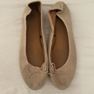 Beige ballerinaskor med rosett från H&M - Säljer ett par klassiska beige ballerinaskor från H&M med rund tå och söt rosettdetalj framtill i mocka material. Skorna har en mjuk ovandel och liten klack, perfekta för en stilren vardagslook.