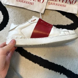 Valentino Open sneakers med röd detalj - Säljer ett par vita Valentino Open sneakers med bred röd läderdetalj över mitten. Skorna har rund tå, platt sula och klassisk Valentino-design. Kommer med originalpåse och låda. Perfekta för dig som gillar stilrena och exklusiva sneakers.