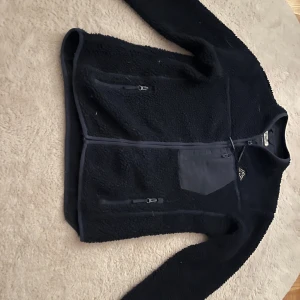 marinblå fleecejacka från Kappa - Mysig marinblå fleecejacka från Kappa med hel dragkedja och två fickor framtill. Jackan har en extra ficka på bröstet med dragkedja och Kappa-logga broderad på bröstet. Perfekt för kyliga dagar. kan finnas spår av hund hår men försöker få bort allt innan den skickas 