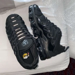 Nike Air Vapormax Plus helsvarta sneakers - Säljer ett par helsvarta Nike Air Vapormax Plus sneakers med futuristisk design och synliga luftbubblor i sulan. Skorna har snörning och en glansig ovandel i syntetmaterial. Perfekta för dig som gillar streetwear och sportig stil.