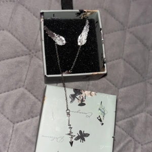 Silverfärgat halsband med vingar och blixt - Unikt örhänge i silverfärg med två detaljerade vingar och ett hängsmycke i form av en blixt. Det är tunn och smycket levereras i en fin ask med fjärilsmotiv. Perfekt för dig som gillar smycken med coola detaljer. Köptes för 699kr (kan även byta mot guld, eller något annat!)