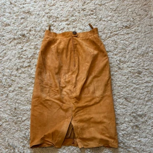 Beige mockakjol med slits - Säljer en beige långkjol i mocka med hög midja och slits framtill. Kjolen har en knapp och dragkedja i midjan samt smala hällor. Perfekt för dig som gillar en stilren och tidlös look.