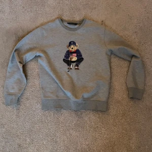 Grå sweatshirt med Björn från Polo Golf Ralph Lauren - Säljer en grå sweatshirt från Polo Golf Ralph Lauren med en broderad björn på framsidan som håller en röd kopp. Tröjan har rund hals och långa ärmar. Perfekt för en avslappnad och stilren look.