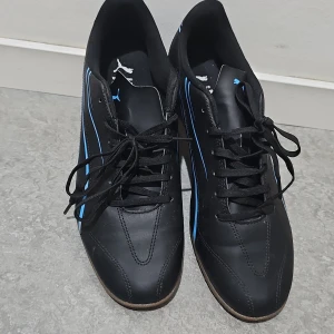 Svarta sneakers från Puma med blå detaljer - Säljer ett par svarta Puma sneakers med blå ränder och detaljer. Skorna har snörning, platt sula och rund tå. Perfekta för dig som gillar sportig stil och vill ha något bekvämt till vardags.