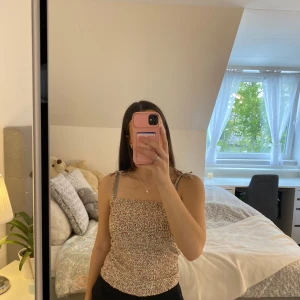 Blommig croppad linnetopp från Vila - Supersöt croppad linnetopp från Vila med smala axelband och blommigt mönster i rött och grönt på beige botten. Perfekt för sommardagar och enkel att matcha med jeans eller kjol.