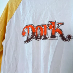 Vit och gul baseball t-shirt med tryck från Vailent Pure - Vit t-shirt med gula raglanärmar och texten 'Dork' i rött och svart på bröstet. Klassisk baseballstil med trekvartsärm. Tillverkad i 100% bomull för en skön och avslappnad känsla.