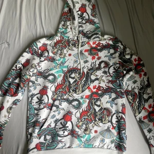 Vit hoodie med drakmönster från Cropp - Säljer en vit hoodie från Cropp i storlek M med färgglatt drakmönster och orientaliska detaljer. Tröjan har huva med dragsko och en stor magficka. Perfekt för dig som vill sticka ut med en unik stil!