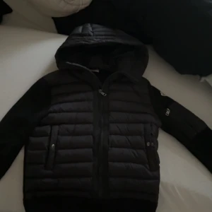 Svart pufferjacka med huva från Moncler - Säljer en svart pufferjacka från Moncler med huva och dragkedja. Jackan har quiltade partier framtill och på ryggen, samt två fickor med dragkedja. Perfekt för kyliga dagar och riktigt snygg streetstil. Storlek S. Säljer eftersom jag ska köpa en moncler väst. Köpte den i februari. Använt den 6-7 gånger då jag ofta använt billigare jackor för att vara rädd om denna! Som tex: peak performance, under armour osv.