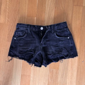 Svarta jeansshorts med fransar - Säljer ett par svarta jeansshorts med fransiga benslut och slitna detaljer framtill. Shortsen har klassisk femficksdesign, bälteshällor och knappgylf. Perfekta för en avslappnad stil.
