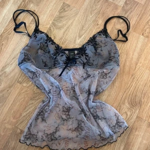 mesh linnetopp - Säljer en transparent mesh linnetopp med svarta spetsdetaljer och änglamotiv. Toppen har tunna axelband och vågig kant nertill. 