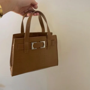 Brun handväska med krokodilstruktur och silverdetalj - Säljer en snygg brun handväska i syntetmaterial med krokodilpräglad yta. Väskan har två handtag och en dekorativ silverfärgad spänndetalj framtill. Perfekt storlek för det viktigaste  och ger en trendig touch till din outfit. Långt band medkommer!