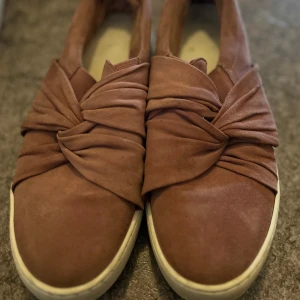 Rosa slip-on skor med knutdetalj - Säljer ett par snygga slip-on skor i rosa mocka med unik knutdetalj framtill. Skorna har vit platt sula och rund tå, vilket ger en stilren och modern look. Perfekta för dig som vill ha något extra till vardagsstilen.