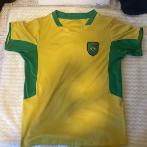 Gul och grön Brasilien-inspirerad fotbollströja - Säljer en gul fotbollströja med gröna detaljer och Brasilien-inspirerat märke på bröstet. Tröjan har korta ärmar och gröna paneler på sidorna samt runt halsen. Perfekt för fotbollsträning eller att visa support för Brasilien.