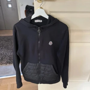 Svart hoodiejacka från Moncler - Säljer en svart hoodiejacka från Moncler med dragkedja och quiltade fickor framtill. Jackan har huva och Moncler-logga på bröstet. Perfekt för en stilren och sportig look. Finns däremot 3 små hål som inte syns vid användning skriv för bilder eller för mer frågor!