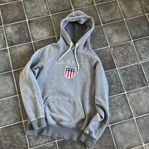 Grå hoodie från GANT med broderad logga - Säljer en grå hoodie från GANT med broderad logga och sköld på bröstet. Tröjan har huva med dragsko och en stor magficka. Klassisk och stilren design med ribbade muddar vid ärmslut och nederkant. Har lite blekta linjer på vissa ställen, se bild 4 och 5. 