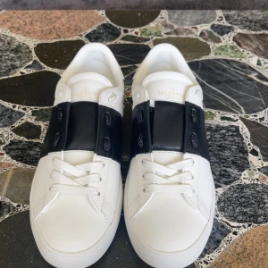 Valentino opens vita svarta .Storlek (42)  - Snygga vita sneakers från Valentino Garavani med bred svart läderdetalj över snörningen och ikoniska nitar runt sulan. Skorna har rund tå och platt sula, perfekt för en stilren och modern look.