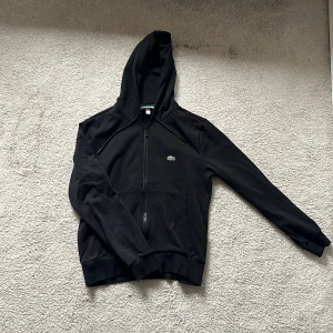 Svart hoodie från Lacoste med dragkedja - Säljer en svart hoodie från Lacoste med dragkedja framtill och klassisk krokodillogga på bröstet. Tröjan har huva med snörning och två fickor framtill. Perfekt för en sportig och stilren look.