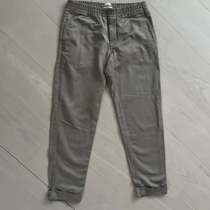 Beige chinos från Filippa K - Snygga beige chinos från Filippa K med elastisk midja och raka ben. Byxorna har fickor i sidorna och bakfickor, samt uppvikta benslut för en avslappnad look. Perfekta till vardags eller när du vill vara lite extra stilren.