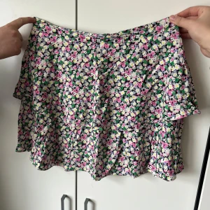 Blommig kortkjol i bomull - Supersöt kortkjol med blommigt mönster i rosa, grön, vit och svart. Kjolen har en lätt utställd form och är perfekt för varma dagar. Bekväm och luftig med mjukt bomullstyg. 