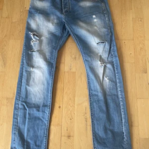 Blå slitna jeans från Burberry - Säljer ett par blå jeans från Burberry med slitningar och ljusa tvättade partier. Klassisk femficksmodell med Burberry-lapp bak i midjan och rutigt tygdetalj på ena bakfickan. Raka ben och normal passform.