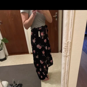 Svart blommig kjol från H&M Divided - Supersnygg svart kjol med rosa och vita blommor från H&M Divided. Kjolen har en omlott-look med slits på sidan och är perfekt för dig som gillar en feminin stil. Längden är mitt emellan kort och lång, vilket gör den lätt att matcha.