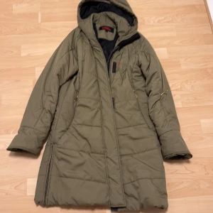 Olivgrön lång dunjacka med huva från Fjällräven - Säljer en olivgrön lång dunjacka från Fjällräven med huva och dragkedja. Jackan har två sidofickor med dragkedja och broderad logga på bröstet. Perfekt för kalla dagar och har en klassisk, stilren design.