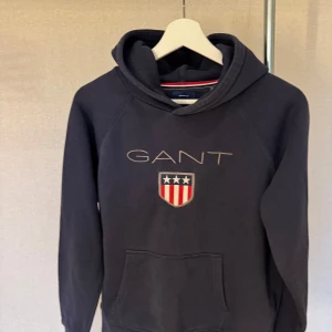 Mörkblå hoodie från GANT med broderad logga - Säljer en mörkblå hoodie från GANT med broderad logga och sköld på bröstet. Tröjan har huva och en stor magficka framtill. Perfekt för dig som gillar klassisk och sportig stil.
