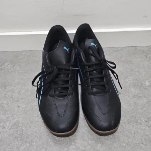 Svarta sneakers från Puma med blå detaljer - Säljer ett par svarta Puma sneakers med blå ränder på sidorna och bruna sulor. Skorna har klassisk snörning och rund tå, perfekt för dig som gillar sportig stil. Puma-logga syns både på plös och insida.