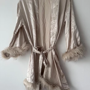 Beige satin kimono med fuskpälsdetaljer - Säljer en beige kimono-inspirerad  morgonrock  i satin med fuskpäls vid ärmslut och nederkant. Jackan har knytband i midjan och en glansig finish som ger en lyxig känsla. Perfekt för dig som vill sticka ut lite extra.