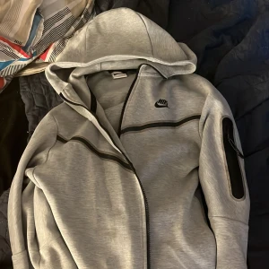 Grå hoodiejacka från Nike - Säljer en grå hoodiejacka från Nike med svarta detaljer och dragkedja. Jackan har lång ärm, huva och en ficka med dragkedja på ena ärmen. Perfekt för träning eller chill. Den funkar också med att bara gå i hemma den är väldigt väldigt skön. Denna klassiska Tech Fleece Windrunner erbjuder polerad värme och komfort, men med reflekterande designdetaljer som lyser upp din look Blixtlåsfickor gör att du säkert kan förvara dina saker i när du är ute ❤️ Priset diskuteras 