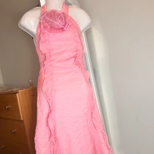 Rosa halterneckklänning med volang och blomdetalj - Supersöt rosa klänning med halterneck och stor tygblomma vid halsen. Klänningen har volangdetaljer längs framsidan och ett lätt rutigt mönster i tyget. Perfekt för somriga tillfällen när du vill sticka ut lite extra.              Längd 78cm