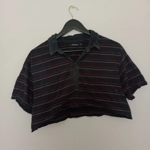 Svart croppad skjorta med ränder - Säljer en svart croppad skjorta från Batistini med röda och vita ränder. Skjortan har krage och korta ärmar, perfekt för en avslappnad stil. Passar dig som gillar unika plagg.