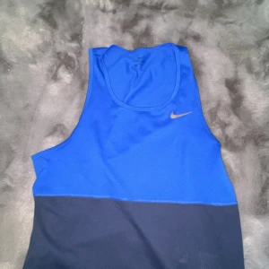 Blå och mörkblå linnetopp från Nike - Säljer en sportig blå och mörkblå linnetopp från Nike med klassisk logga på bröstet. Toppen är ärmlös och tillverkad i ett lätt och ventilerande syntetmaterial, perfekt för träning.