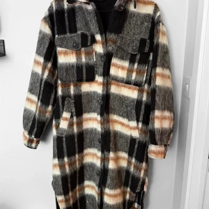 Rutig lång jacka från Hains - Säljer en lång, rutig jacka från Hains i svart, grått, beige och vitt. Jackan har stora bröstfickor med knappar, knäppning framtill och klassisk krage. Materialet är mjukt och borstat, perfekt för kyligare dagar.