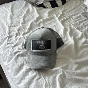 Grå keps med bilmotiv från ENZO - Snygg grå keps med mesh baktill och ett stort svartvitt motiv av en bil framtill. Justerbar passform med plastspänne. Perfekt för en avslappnad stil och streetwear. Aldrig andvänd. Vill bara bli av men den tveka inte på och lägga prisförslag!