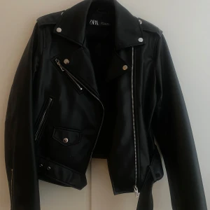 Svart skinnjacka från Zara - Säljer en svart skinnjacka från Zara med klassisk bikerstil. Jackan har silvriga dragkedjor, knappar och detaljer samt bälte i midjan. Snygg krage och flera fickor framtill. Perfekt för dig som vill ha en edgy look.