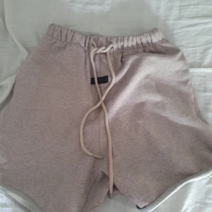 Beige shorts från Fear of God Essentials - Säljer ett par snygga beige shorts från Fear of God Essentials. De har elastisk midja med snörning och är tillverkade i mjuk bomull. Perfekta för en avslappnad stil.