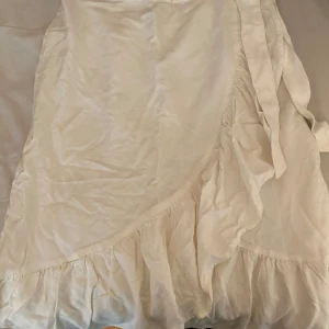 Vit omlottkjol från Pimkie - Säljer en vit omlottkjol från Pimkie med volangdetalj längs nederkanten. Kjolen har knytband i midjan och är perfekt för en fräsch och somrig look. Lätt och luftig modell som passar till många olika stilar. Skulle gissa på att storleken är runt xs/s men vet inte helt säkert! 