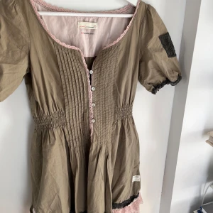 Beige klänning med spets från Odd Molly - Supersöt beige klänning från Odd Molly med rosa spetsdetaljer och svarta spetskanter. Knappar framtill, kort ärm och smock i midjan. Fina veckade detaljer på bröstet och broderad text på ryggen. Perfekt för dig som gillar unika detaljer!